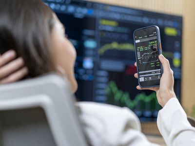 Woman looking at a stock chart on a smartphone with another larger screen with stock charts on it in the background