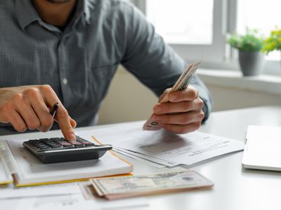 Man using calculator while holding money at desk with files, a computer, and an open notebook