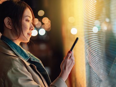 A person looks at a smartphone while illuminated by a digital display at night.