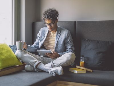 Young man sitting at home on his couch, looking intently at a table with a coffee cup in his other hand.