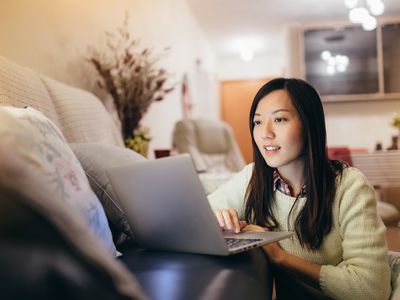 A woman wearing a white sweater sits on the ground facing a sofa looking at a laptop that is on the sofa