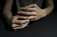 A dark background surrounding a woman's hands close up with one hand pulling off a wedding ring. 