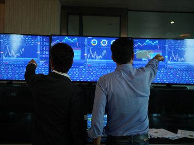 Two people pointing at screens with stock information while standing.