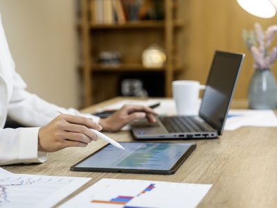 A close-up of a person's hands using a stylus pen to point to graphs on a tablet, while sitting in front of a laptop.