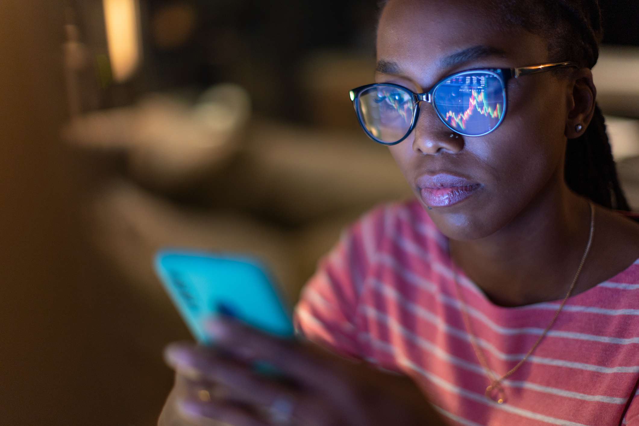 A woman wearing glasses holds a smartphone, and the screen of the phone is pointed to her face but is reflected in her glasses. It displays stock chart data. 