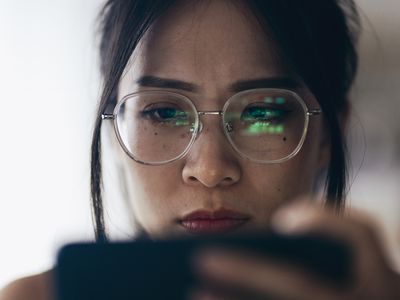 A woman holding a smartphone that is being reflected in her glasses with green charts and/or words