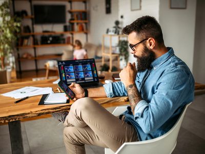 Man with a beard sits at a kitchen table holding a smartphone in front of a laptop and both screens are showing stock charts. A child sits in the background.