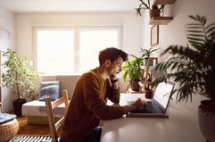 A man with a brown shirt on sits at a table in a living room with a laptop surrounded by natural light and several house plants