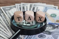 Cube wooden blocks with word TAX on a magnifying glass on US dollar banknotes on a wood table.