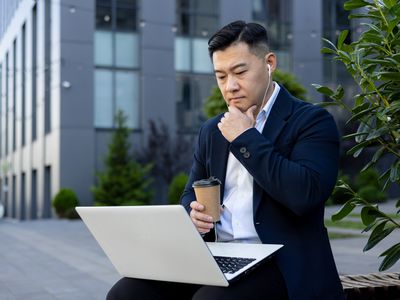 Man sitting on a bench outside an office building, looking at a laptop.