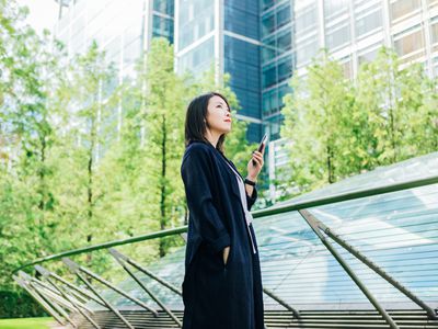 Young asian woman holding her smartphone, standing against corporate building surrounded by trees.