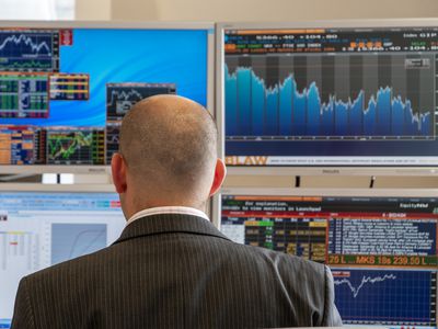 A man sits with his back to the camera at a desk with four computer screens displaying various charts.