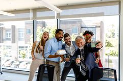 Group of people smiling brightly. One person is holding a cake as one of the professional people takes a selfie of them all.