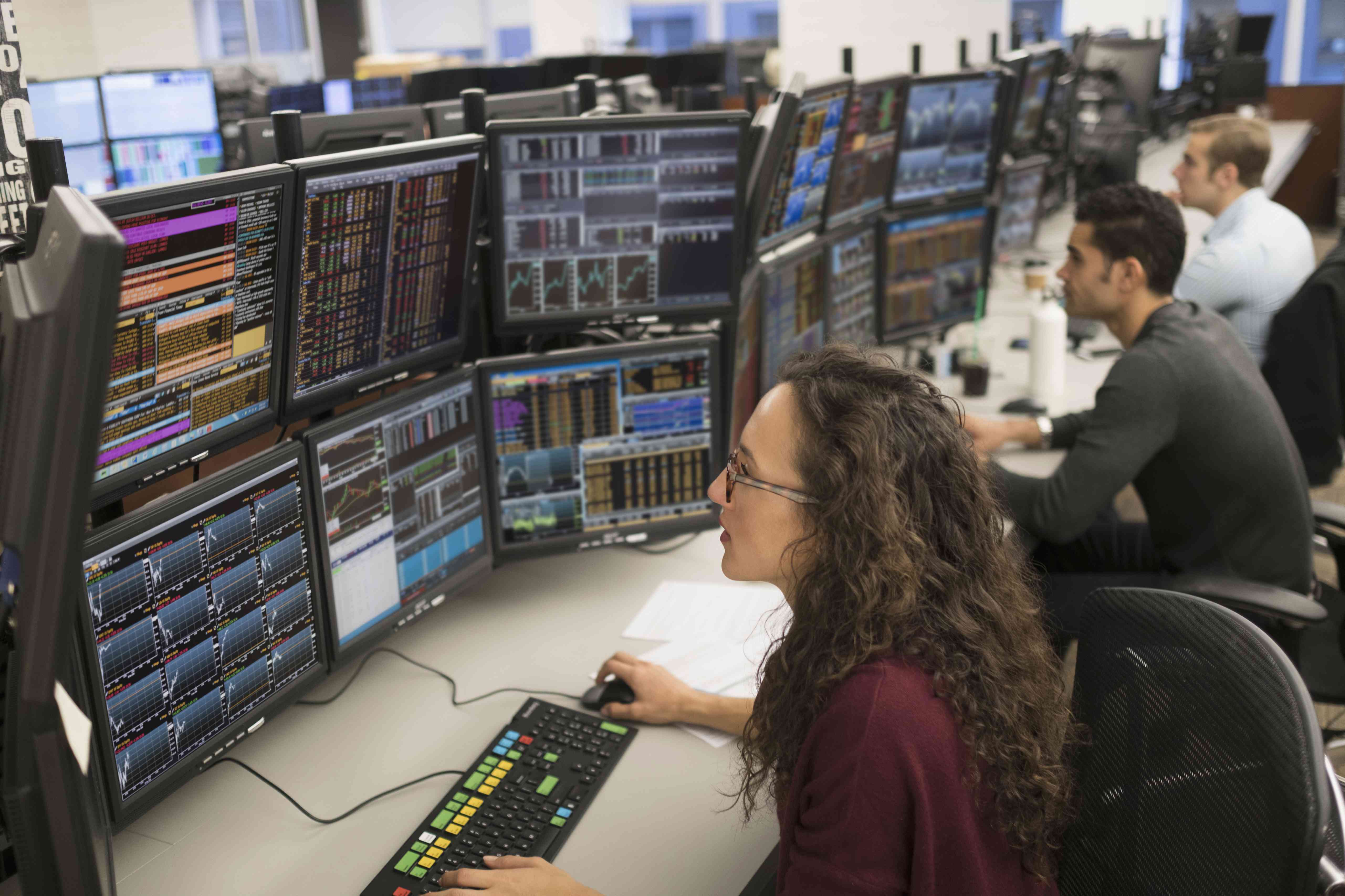 A row of traders at trading desks in a brokerage use computer systems with multiple screens to enter orders for clients.