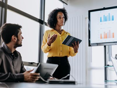 A person leads a discussion with bar charts on a screen.