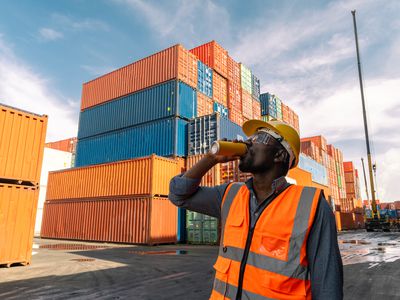 Worker drinks from a can while being surrounded by shipping containers.