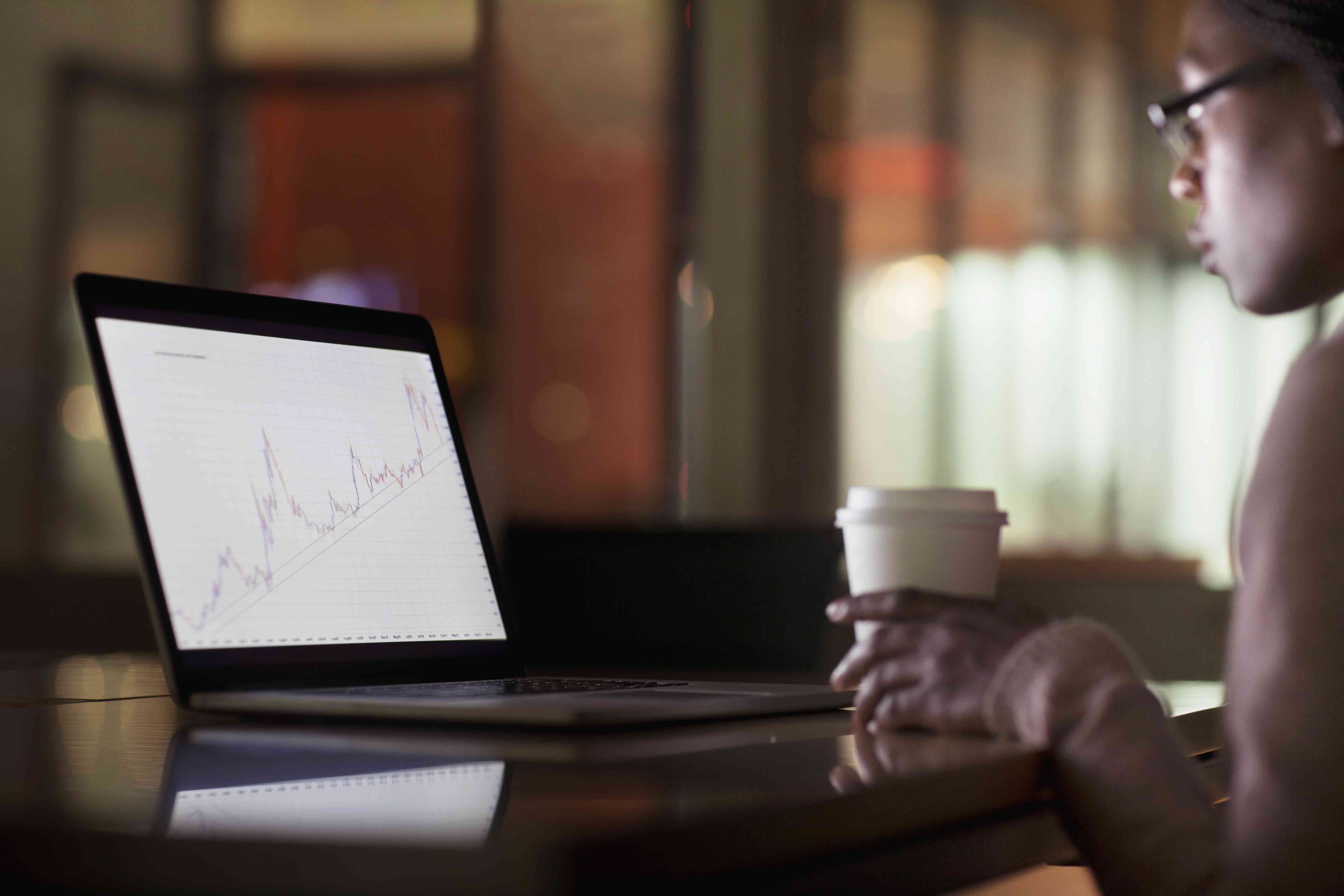 A person looks at a stock chart on a laptop while holding a coffee cup