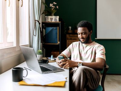 Young adult with beard reviews stock investments on phone while sitting next to a window.