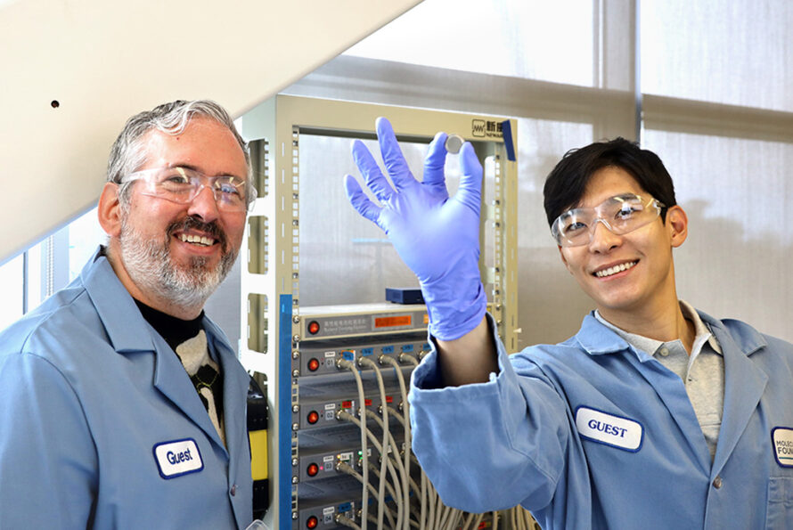 Two smiling people in blue lab coats and safety glasses. The person on the right has their arm raised in the center of the frame presenting a nickel-sized sample.