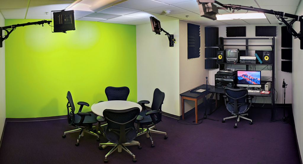 Large room with a green wall, table and chairs, studio lights, and a computer workstation with audio and video equipment