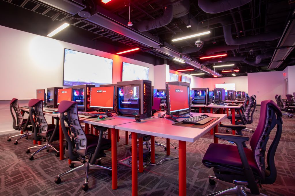 Large room with red lighting, gaming chairs, and tables topped with high-end PCs and monitors