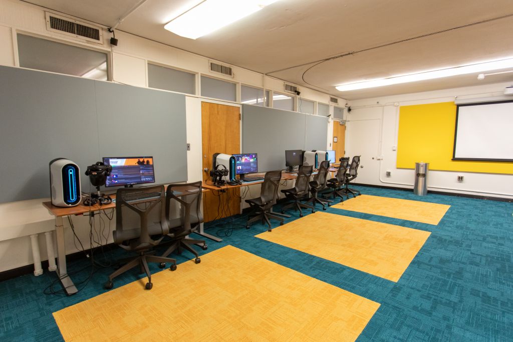 The room features a series of VR-equipped computer workstations along one wall, and a projector screen on the opposite wall, indicating a training or presentation area.