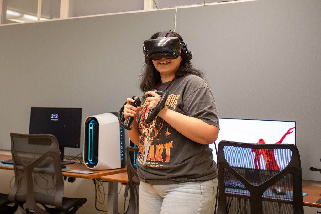 A participant is engaged in a virtual reality experience in a well-equipped room. She stands in front of a desk setup with a high-performance desktop PC and a monitor displaying a red abstract figure. The room features modern design elements with ergonomic chairs and clean lines.