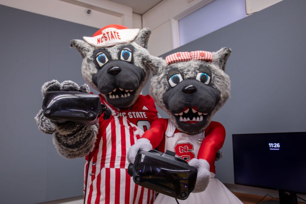 Both mascots hold VR headsets and smile, promoting the studio's gaming and VR facilities. Mr. Wuf is in a red and white striped outfit, while Ms. Wuf wears a cheerleading dress, both representing NC State.