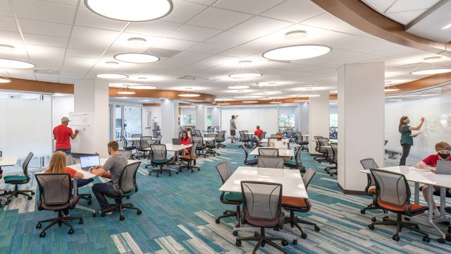 Large, well lit room with lots of tables and chairs. Students in masks are studying and drawing diagrams on whiteboards