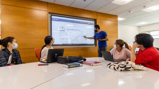 A person draws a graph on a very large touchscreen that is mounted to a wall. Four other people look on in interest