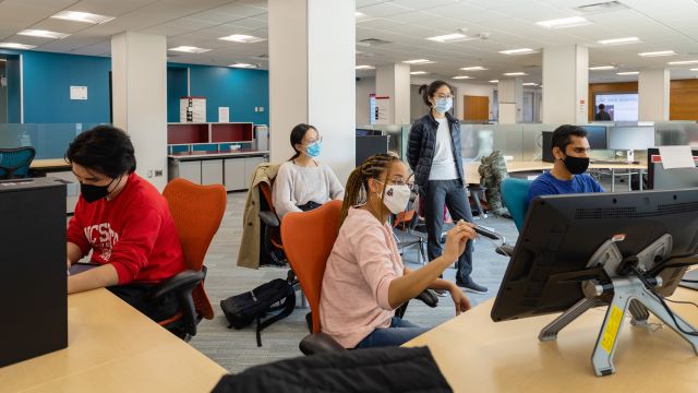 A group of people sit in colorful chairs in the computer lab. One person holds a stylus up to a large tablet.