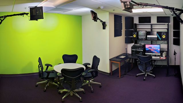 Large room with a green wall, table and chairs, studio lights, and a computer workstation with audio and video equipment