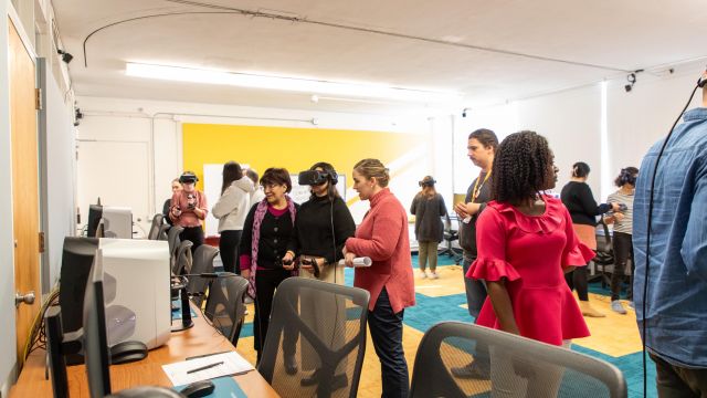 The room is bustling with activity as a group of people explores VR experiences. Several individuals wear VR headsets, interacting with virtual content using controllers. Others watch or wait their turn, engaged in conversation.