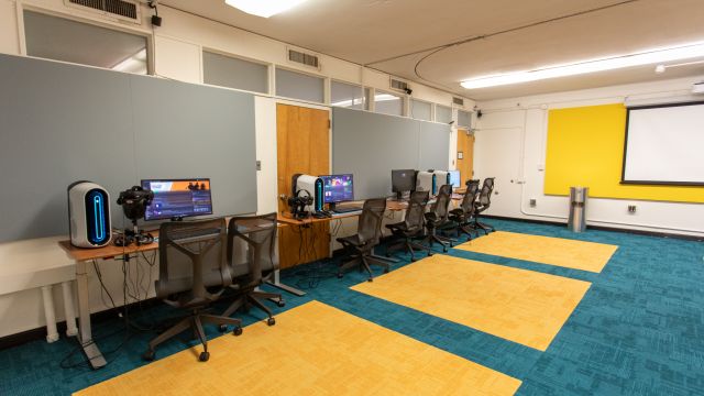 The room features a series of VR-equipped computer workstations along one wall, and a projector screen on the opposite wall, indicating a training or presentation area.