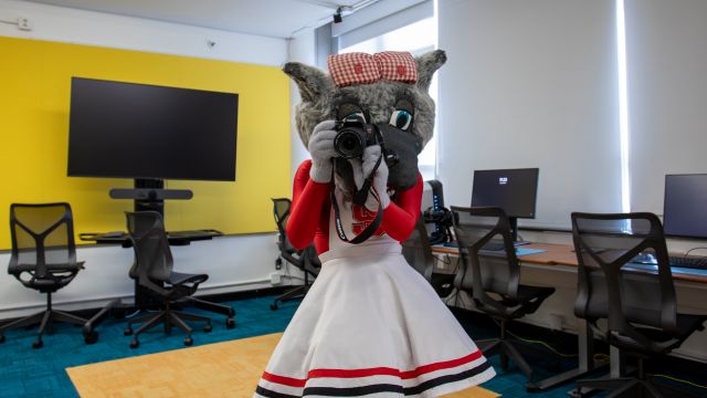 Dressed in a cheerleading outfit with NC State branding, Ms. Wuf holds a DSLR camera, demonstrating the VR studio activities.