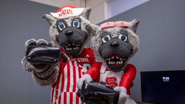 Both mascots hold VR headsets and smile, promoting the studio's gaming and VR facilities. Mr. Wuf is in a red and white striped outfit, while Ms. Wuf wears a cheerleading dress, both representing NC State.