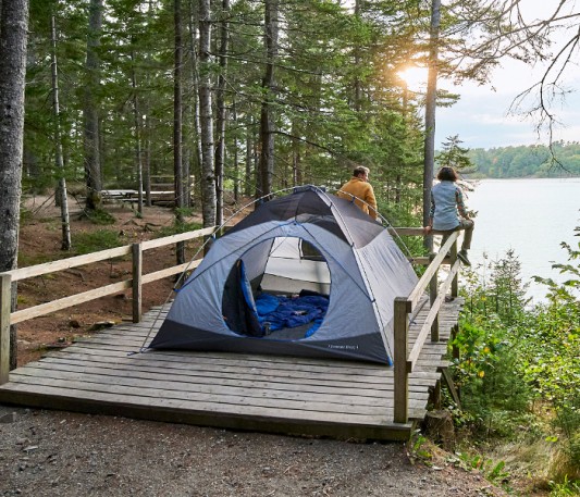  Two people sitting on a fence looking out at the water, their tent pitched behind them.