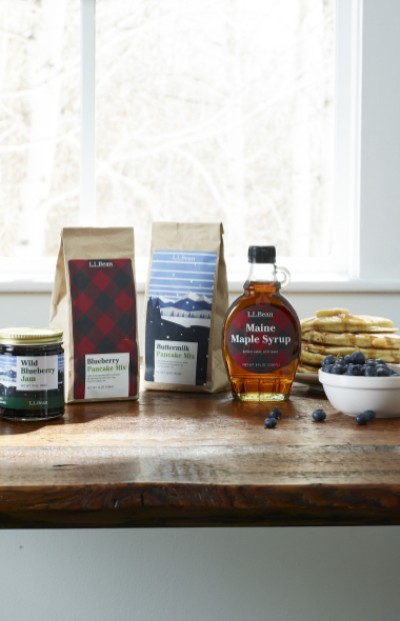 Breakfast items, including maple syrup and pancake mix, are arranged on a wooden table with a window in the background.