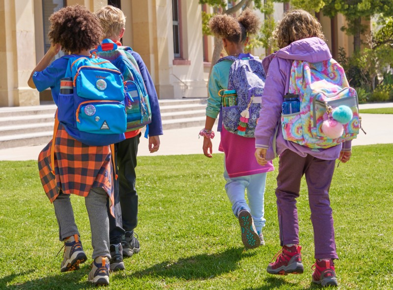 4 kids walking toward their school wearing school backpacks.