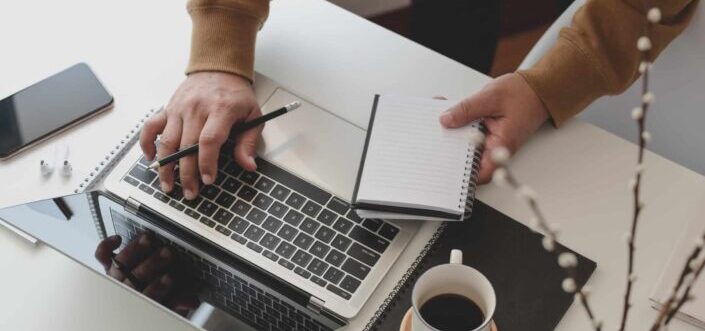 Hand typing on a MacBook while holding a notebook on the other hand