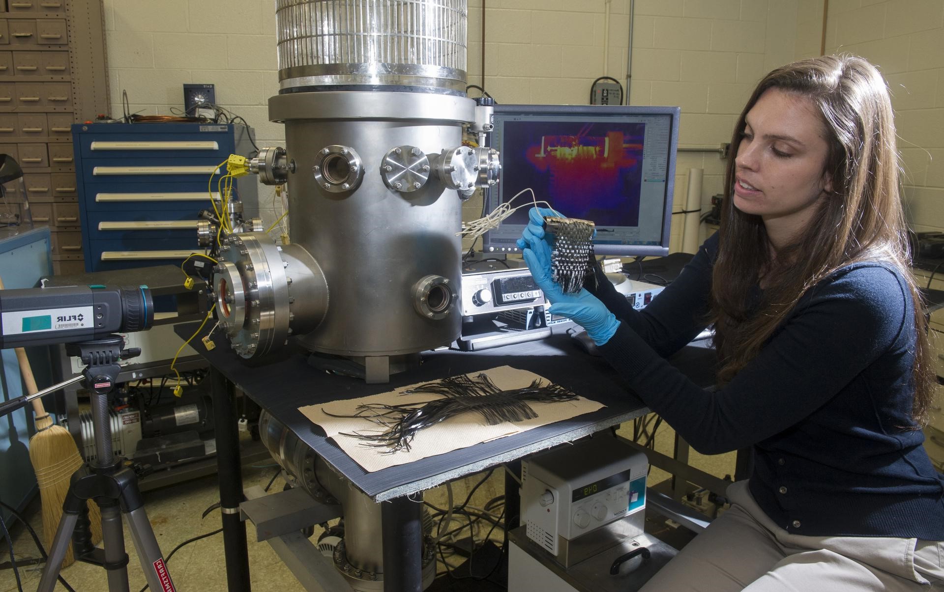 Female sitting in front of a desk, a screen, and a machine, showing something metal.