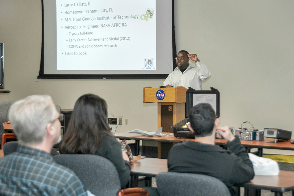 Speaker stands at podium in front of several people in the audience. A screen, showing a presentation slide, is on the wall behind the speaker.