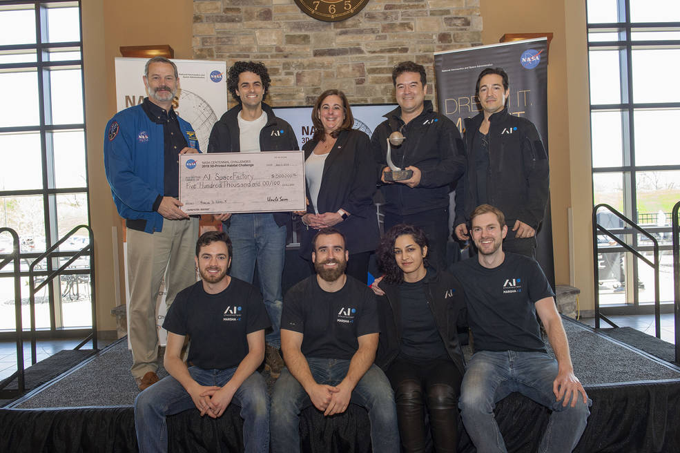 Members of AI. SpaceFactory receive a large check as an award for winning the final round of NASA's 3D Printed Habitat Challenge. Five people stand in the back and four people sit in the front on a stage, with the check being held by a man in a blue NASA jacket on the far left and a member of AI. SpaceFactory holding the check next to the man in the blue jacket. All nine people smile brightly.