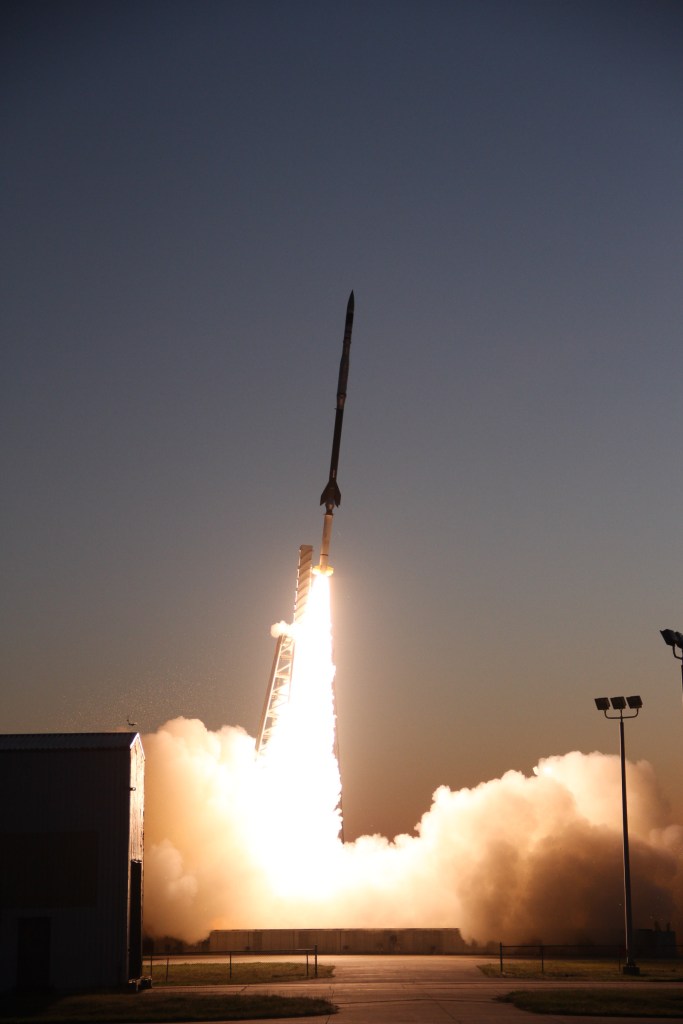 A sounding rocket in mid-launch off the launch pad with a bright white plume the spreads out underneath the rocket when it hits the ground. The rocket is white on the bottom and darker up to the top.