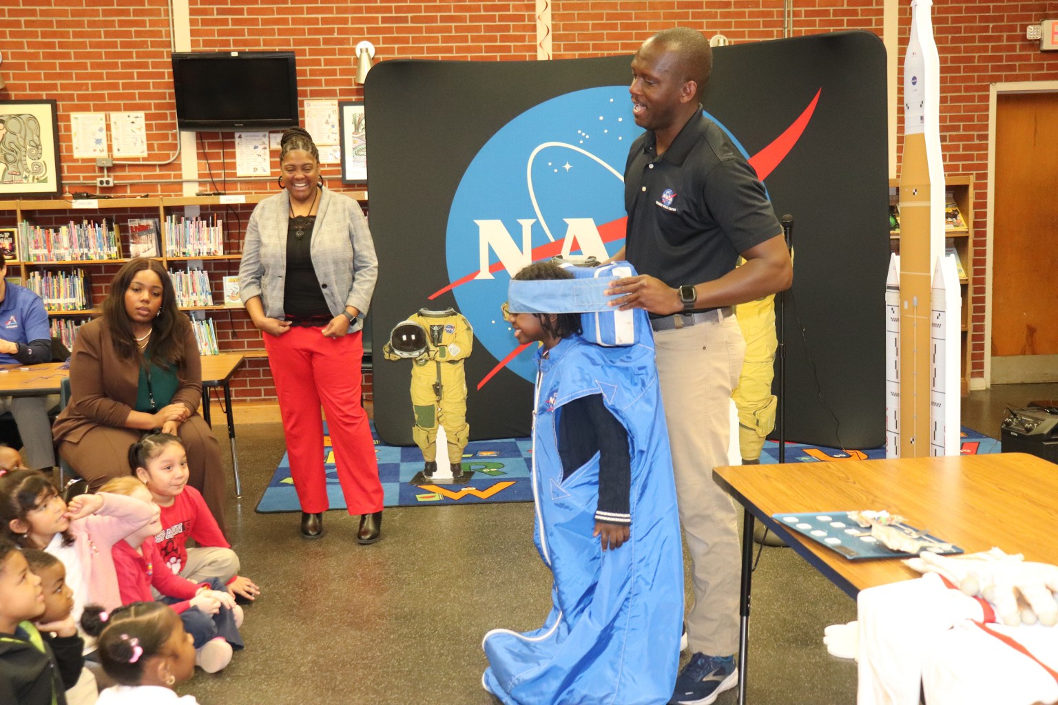 Man and young male student demonstrate how to sleep in space to a group of elementary students