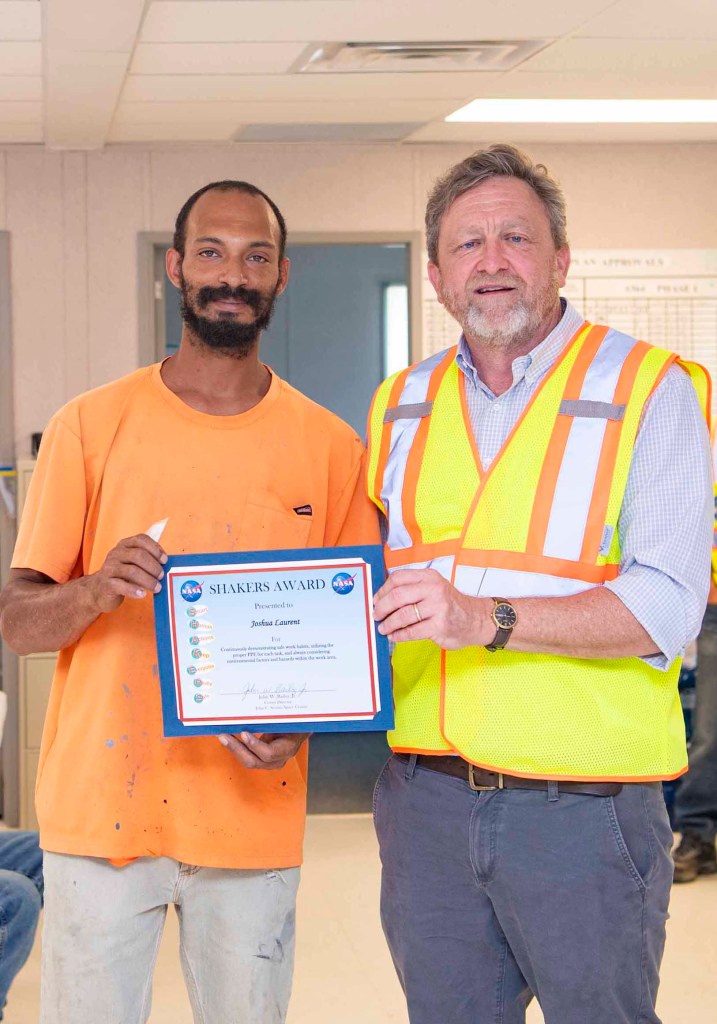 Rodney McKellip, wearing a yellow and orange safety vest, presents a SHAKERS Award to Joshua Laurent, wearing the orange tee shirt