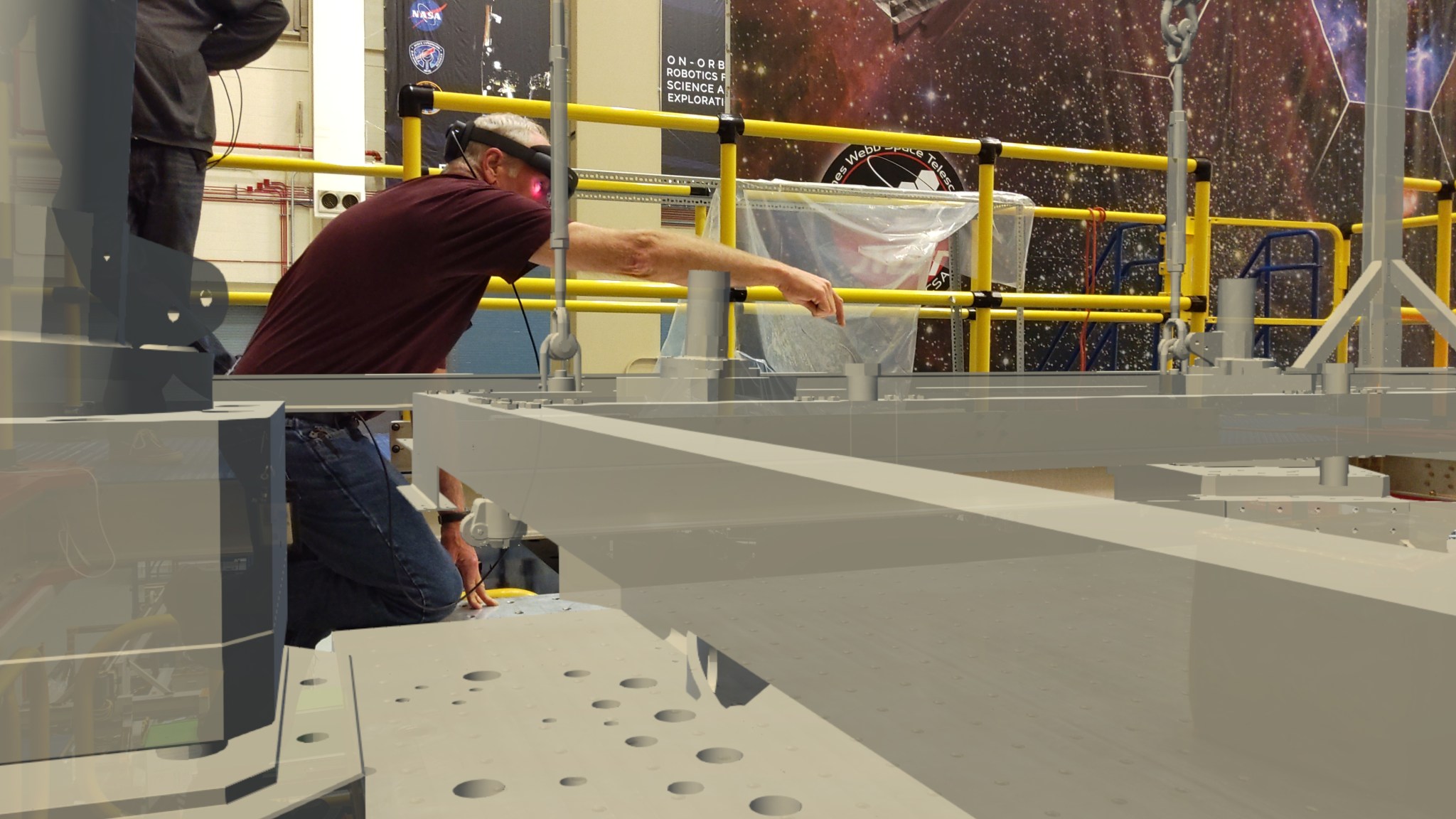 a translucent computer projection of scaffolding is projected into the real environment of a workshop while a person with an AR headset in the background gestures toward a part of the projection