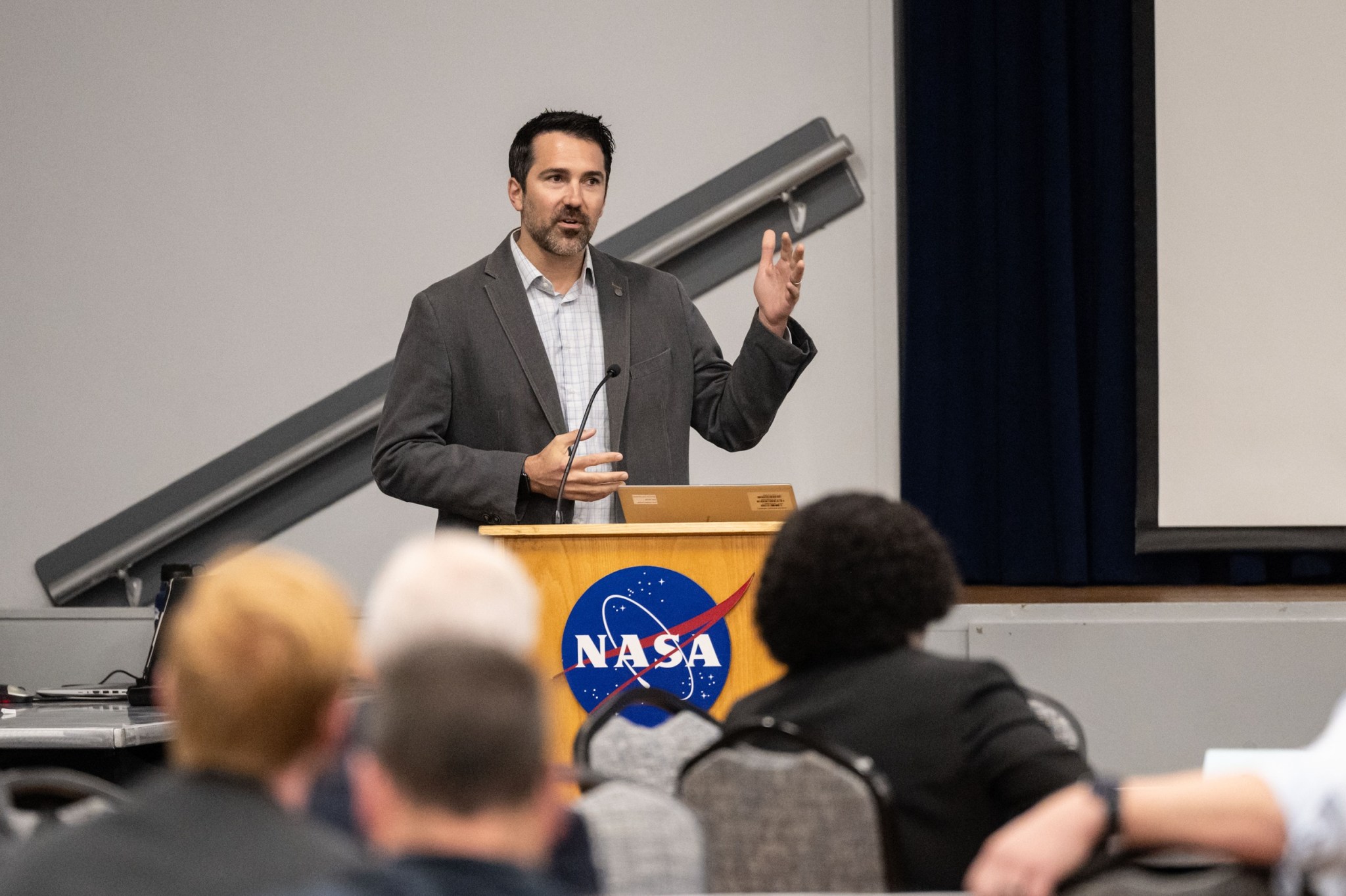 A man wearing a gray blazer stands at a podium with the NASA meatball logo attached to the front. The man is gesturing with his hands as he speaks to an audience.