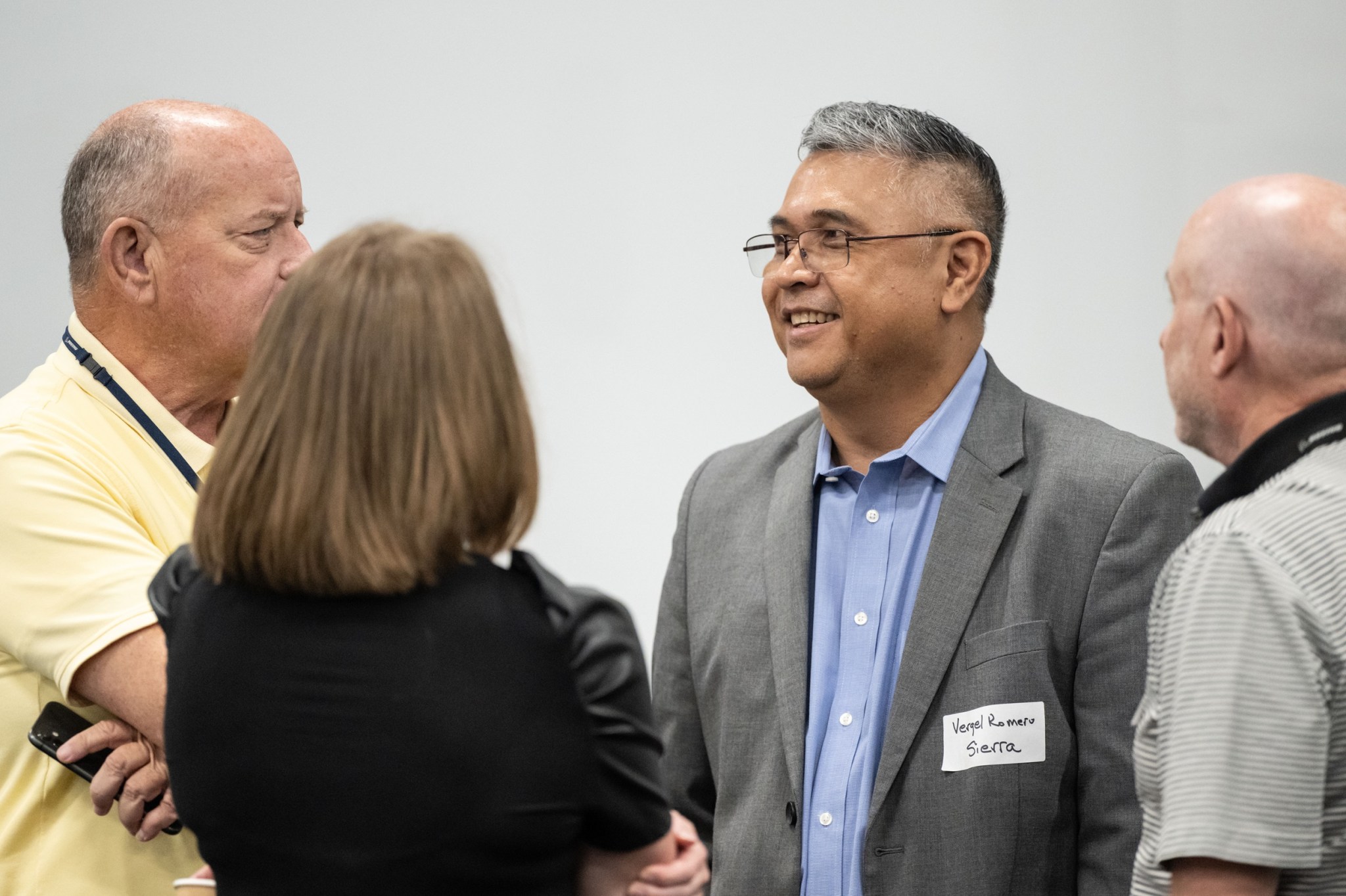 A group of four people are speaking to each other. There are three men and a woman. The man in the center is wearing a blue button-down shirt with a gray blazer and glasses and smiling at the other people. 