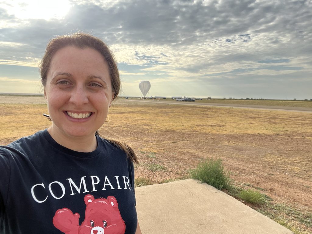 Regina Caputo smiles at the camera in a selfie that captures her head and shoulders. Her brown hair is tied back in a ponytail, and she is wearing a navy-blue tee-shirt which reads “COMPAIR.” Behind Regina is a large open field covered in patches of light brown and yellow grass. The sky is a hazy gray-blue and is covered in dark gray clouds that are thick in certain places but patchy. In the far distance behind Regina, nearing the horizon, is a large space-balloon. The balloon resembles a gray upside-down teardrop, the tip of which just touches the ground.